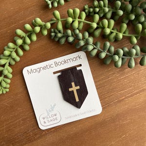 May include: A magnetic bookmark with a dark brown leather banner and a gold cross. The bookmark is attached to a white card with the text "Magnetic Bookmark" and the brand name "Willow & Sage". The words "handmade in South Dakota" are also visible.