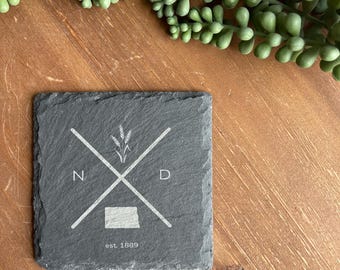 May include: A square, dark gray slate coaster with a white graphic design. The design includes crossed lines, wheat, the letters "N" and "D", a state outline, and the text "est. 1889". The coaster is on a wooden surface with green plant accents.
