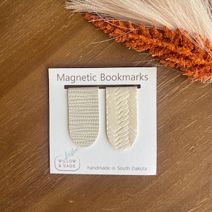 May include: Two white leather magnetic bookmarks with a textured pattern. The bookmarks are on a white card with the text "Magnetic Bookmarks" and "handmade in South Dakota".