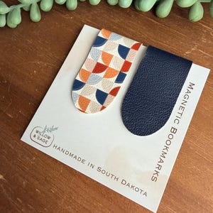 May include: Two magnetic bookmarks attached to a white display card labeled Willow & Sage Handmade in South Dakota. One bookmark features a geometric pattern of orange, navy blue, and cream semicircles, while the other is solid navy blue.