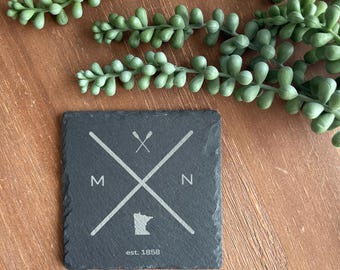 May include: Black slate coaster with a white outline of the state of Minnesota, two crossed paddles, and the letters "MN" with "est. 1858" below.