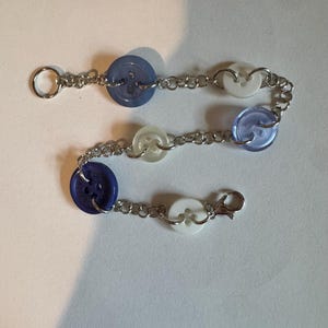 May include: A handmade bracelet featuring a series of blue and white buttons connected by a silver chain. The bracelet has a lobster clasp closure and a small ring at the other end. The buttons are in shades of light and dark blue and white.