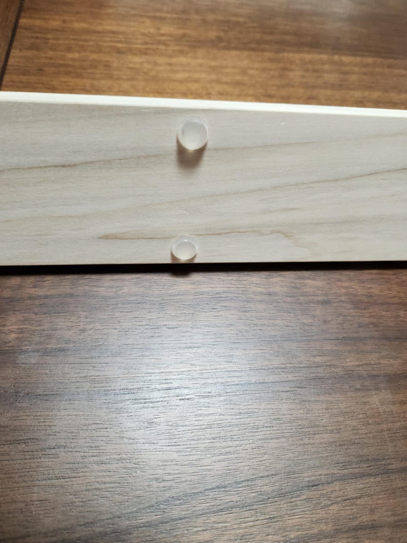 May include: Two clear plastic bumpers are attached to a light wood surface. The wood surface is likely part of a piece of furniture.