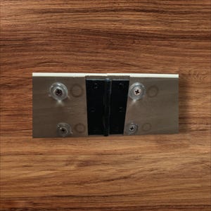 May include: A black metal hinge with four clear plastic screw covers. The hinge is attached to a silver metal plate.