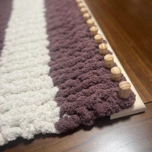 May include: A close-up of a knitted blanket with white and purple stripes. The blanket is being woven on a loom with wooden pegs.