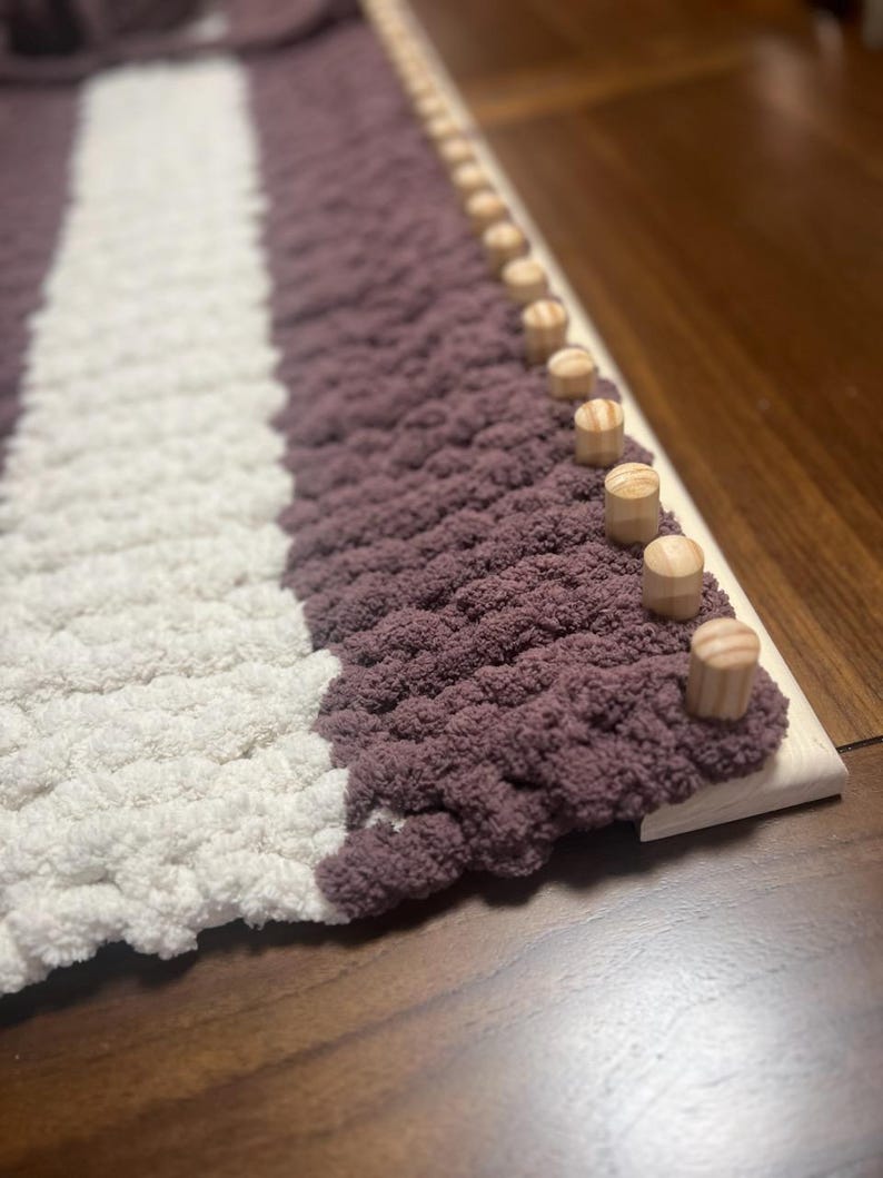 May include: A close-up of a knitted blanket with white and purple stripes. The blanket is being woven on a loom with wooden pegs.