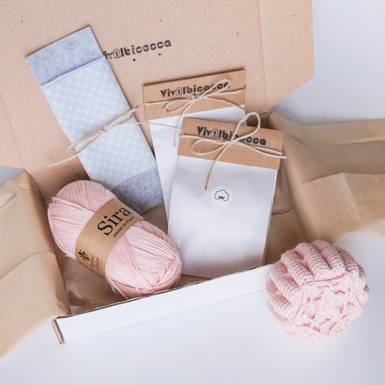 Pu&ograve; includere: Una scatola di cartone marrone con un gomitolo di filato rosa, un tessuto a fantasia blu e bianca e due sacchetti di carta bianca con la scritta "Vivalbico.ca" stampata su di essi.