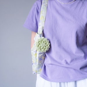 Pu&ograve; includere: Una maglietta color lavanda con una borsa a stampa floreale portata a tracolla. La borsa ha un fiore di uncinetto verde attaccato.