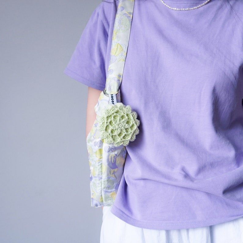 Pu&ograve; includere: Una maglietta color lavanda con una borsa a stampa floreale portata a tracolla. La borsa ha un fiore di uncinetto verde attaccato.