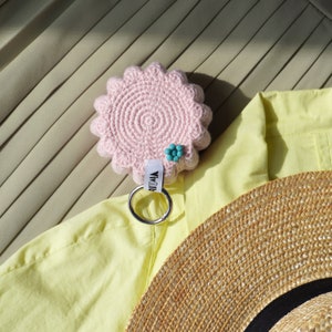 Pu&ograve; includere: Un portachiavi rosa lavorato a uncinetto con un ciondolo a forma di fiore e un anello portachiavi argentato. Il portachiavi &egrave; su uno sfondo di tessuto giallo con un cappello di paglia sullo sfondo.