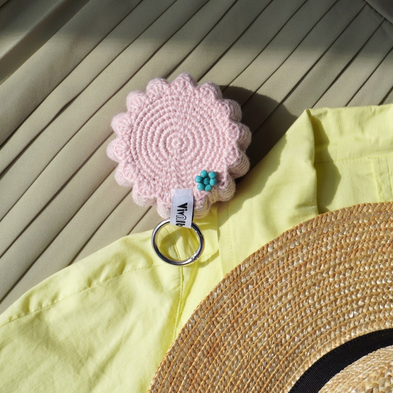 Pu&ograve; includere: Un portachiavi rosa lavorato a uncinetto con un ciondolo a forma di fiore e un anello portachiavi argentato. Il portachiavi &egrave; su uno sfondo di tessuto giallo con un cappello di paglia sullo sfondo.