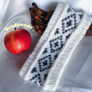 May include: White knit headband with a black and white geometric pattern. The headband is displayed on a white surface with a red apple and a wooden hand.