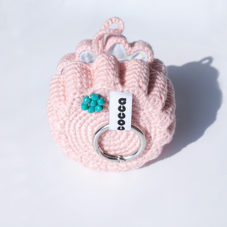 Pu&ograve; includere: Una pochette rosa in crochet con un'etichetta bianca che dice "COCCA" e un abbellimento di perline turchesi. La pochette ha un anello di metallo argentato.