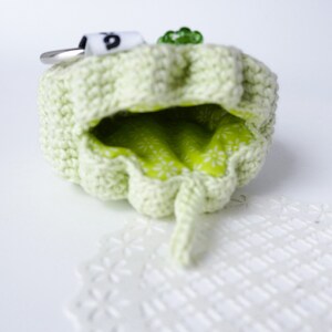 Pu&ograve; includere: Una pochette in crochet verde chiaro con una fodera in tessuto verde e un piccolo portachiavi argentato attaccato. La pochette &egrave; aperta e la fodera &egrave; visibile.
