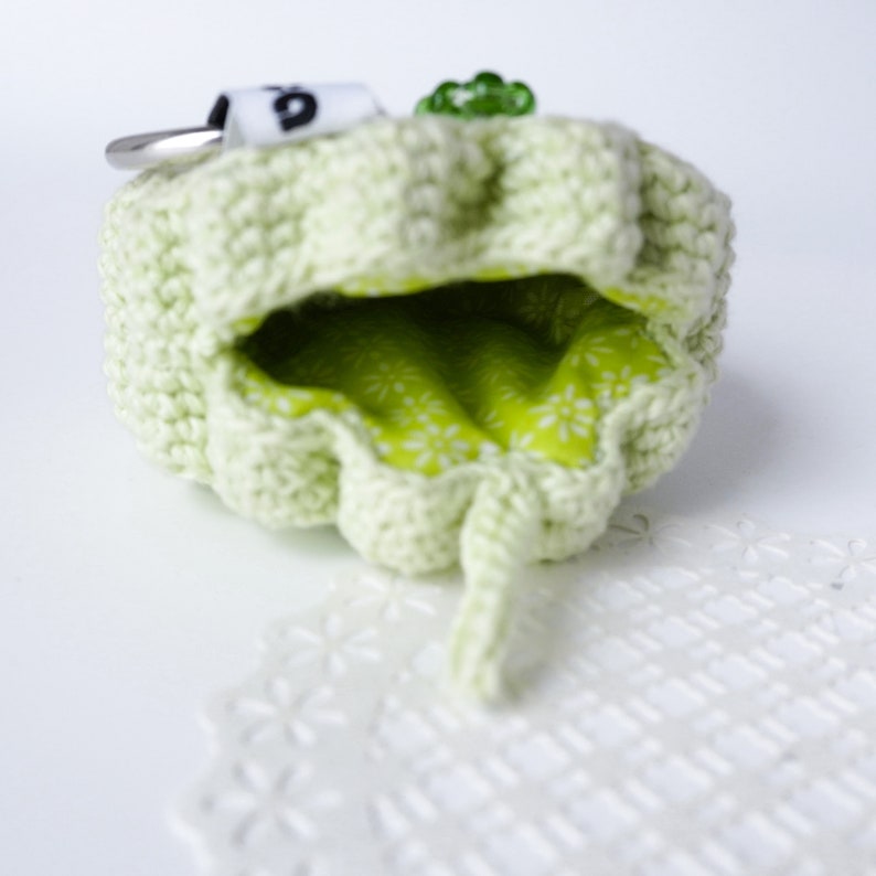 Pu&ograve; includere: Una pochette in crochet verde chiaro con una fodera in tessuto verde e un piccolo portachiavi argentato attaccato. La pochette &egrave; aperta e la fodera &egrave; visibile.