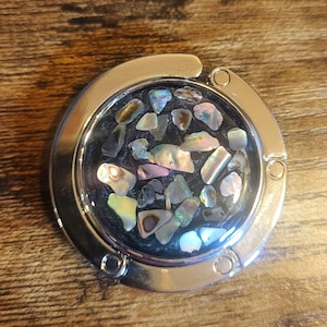 May include: A silver-toned purse hook with a circular design. The center features a mosaic of iridescent, irregularly shaped pieces, showcasing colors like blue, green, and pink. The hook is designed to hang from a table.