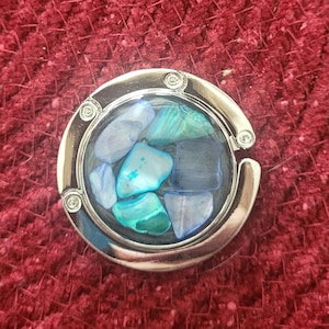 May include: A silver-toned metal purse hook with a round, clear, glass-like insert. The insert is filled with small, blue, green, and white pieces of iridescent material.