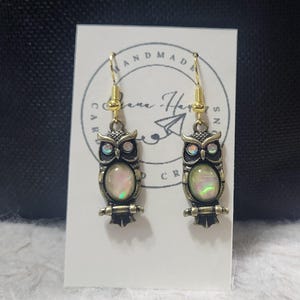 May include: A pair of owl-shaped earrings with gold-colored hooks. The earrings have a bronze-colored frame with iridescent, oval-shaped centers. The earrings are displayed on a white card with the words "HANDMADE" and "Care Haven Creations".
