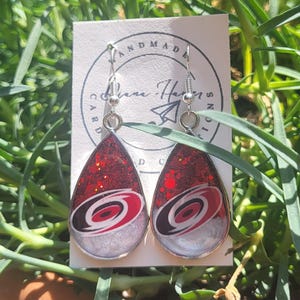 May include: Teardrop-shaped earrings with a red glitter top and a silver base. Each earring features a black, white, and red logo. The earrings are displayed on a white card with the words "HANDMADE" and a signature.