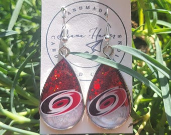 Puede incluir: Pendientes en forma de lágrima con la parte superior de purpurina roja y base plateada. Cada pendiente presenta un logotipo negro, blanco y rojo. Los pendientes se muestran en una tarjeta blanca con las palabras "HANDMADE" y una firma.