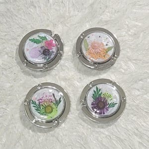 May include: Four silver-toned metal purse hooks with a round, clear, white background. Each hook features a different pressed flower design with a variety of colors, including pink, purple, yellow, and green.