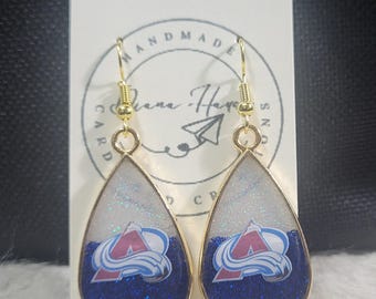 May include: Teardrop-shaped earrings with a gold-colored frame. The earrings feature a blue and white glitter design with the Colorado Avalanche logo. The earrings are hanging in front of a white card with the words "HANDMADE" and "Diane Hayes Designs".