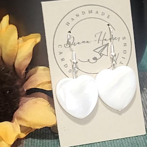 May include: A pair of white heart-shaped earrings with silver hooks. The earrings are displayed on a card with the text "HANDMADE" and a signature. A sunflower is visible in the background.