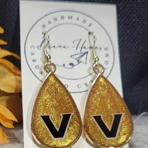 May include: Gold teardrop earrings with a glittery finish and a black "V" design. The earrings have gold-colored hooks and a gold border. The background includes a card that says "HANDMADE CARDS & CRAFTS".