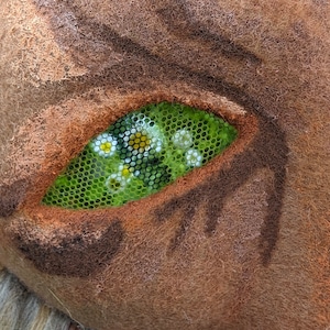 May include: Close-up of a brown, furry creature with a green eye. The eye is made of mesh and has small white flowers inside.