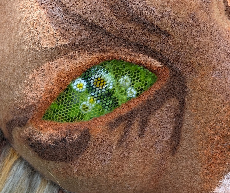 May include: Close-up of a brown, furry creature with a green eye. The eye is made of mesh and has small white flowers inside.