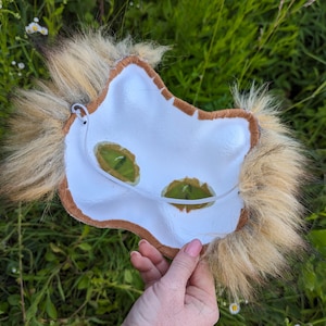 May include: The back of a brown and white furry animal mask with a white felt base and brown felt trim. The mask has two green mesh eye holes and a white elastic strap.