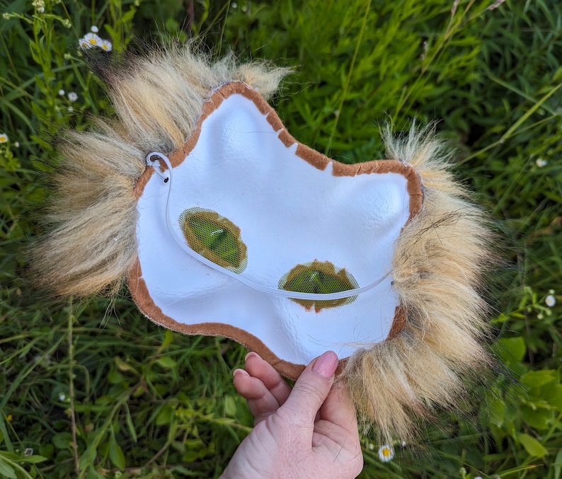 May include: The back of a brown and white furry animal mask with a white felt base and brown felt trim. The mask has two green mesh eye holes and a white elastic strap.