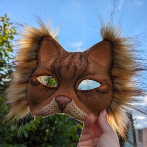 May include: A brown and tan cat mask with fur trim around the edges. The mask has a realistic cat face with black eyes and a pink nose.