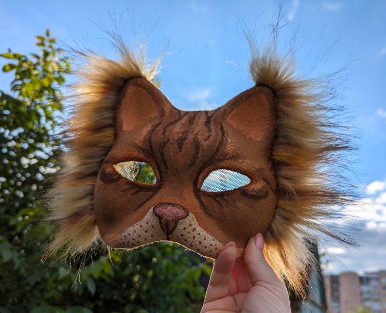 May include: A brown and tan cat mask with fur trim around the edges. The mask has a realistic cat face with black eyes and a pink nose.