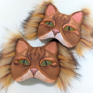 May include: Two brown cat masks with fluffy fur trim. The masks have green eyes, pink noses, and brown and white fur details.
