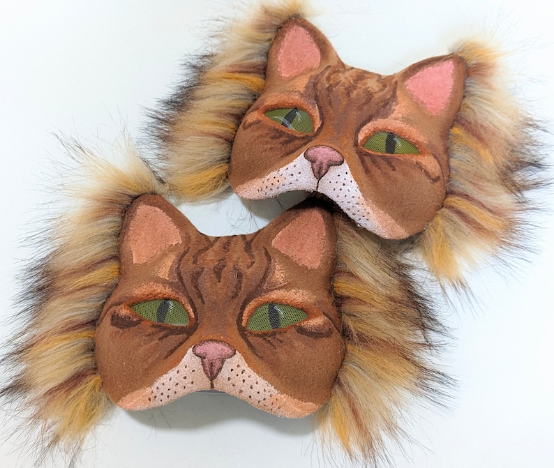 May include: Two brown cat masks with fluffy fur trim. The masks have green eyes, pink noses, and brown and white fur details.