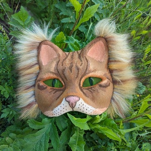 May include: A brown and tan cat mask with green eyes and a fluffy white and brown mane. The mask is made of felt and has a realistic look.