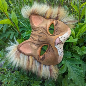 May include: A brown and tan cat mask with green eyes and a furry mane. The mask is made of felt and has a realistic look.