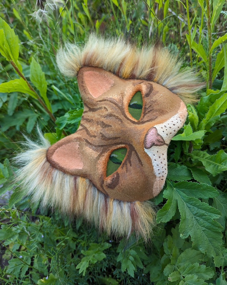 May include: A brown and tan cat mask with green eyes and a furry mane. The mask is made of felt and has a realistic look.