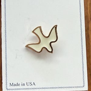 May include: A gold-coloured dove pin with a white enamel fill, presented on a white card with a blue border. The card reads "Made in USA". The pin is a stylised representation of a dove, a symbol of peace.
