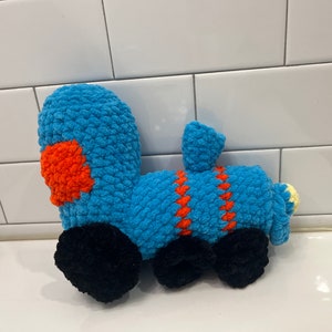 May include: A handmade blue crochet train toy with orange and black accents. The toy train features a rounded body, a chimney, and black wheels. The train is a soft, plush toy.