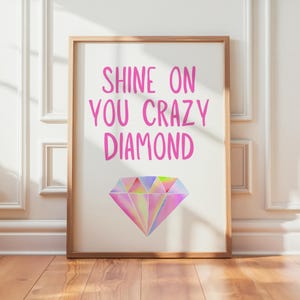 May include: A framed art print with the words "SHINE ON YOU CRAZY DIAMOND" in pink script above a colorful, geometric diamond illustration. The artwork is displayed on a white background with a wooden frame, set against a white wall and wooden floor.