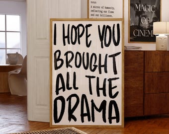 May include: A framed art print with the text "I HOPE YOU BROUGHT ALL THE DRAMA" in bold black letters. The print is displayed on a wooden floor, with other framed art pieces visible in the background.