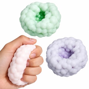Pode incluir: Três brinquedos sensoriais circulares em forma de toro com uma textura irregular semelhante a bolhas. Um é verde-claro, um é violeta-claro e um é rosa-claro, que está sendo apertado por uma mão. Os brinquedos têm um centro oco e flexível.