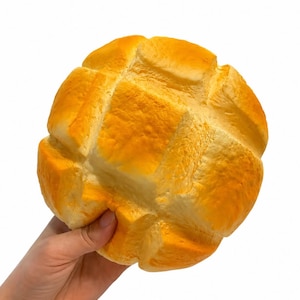 May include: A round, golden-brown squishy toy shaped like a melon pan bread roll. The surface features a distinct grid-like pattern of deep indentations across the top, with a textured, porous appearance that mimics the look of baked crust.