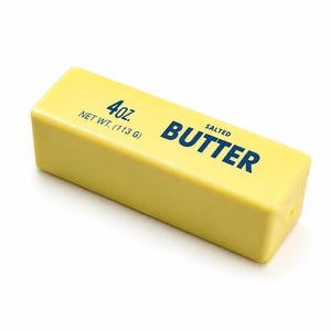 Könnte beinhalten: Ein rechteckiger Riegel gesalzene Butter in einer hellgelben Verpackung. Der Text auf der Oberseite lautet 4oz. NET WT. (113 G) und SALTED BUTTER in fetter, dunkelblauer Schrift. Die Butter ist schräg vor einem weißen Hintergrund platziert.