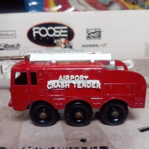 May include: A red toy fire truck with the words "AIRPORT CRASH TENDER" in white lettering. The truck has six black wheels and a white ladder on top. The toy is a miniature replica of a fire truck.