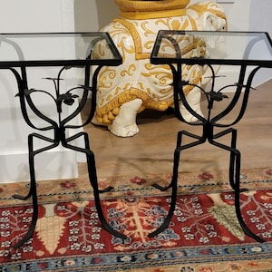 May include: Two black metal side tables with clear glass tops. The tables have a decorative floral design and curved legs. The tables are on a patterned rug. The tables are approximately 24 inches tall.