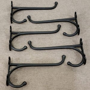 May include: Four black metal plant hooks with a curved design. Each hook has a decorative backplate and a curved arm ending in a rounded tip. The hooks are designed for hanging plants or other items.
