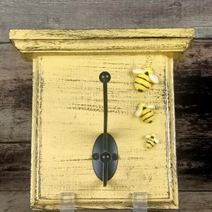 May include: A yellow wooden wall hook with a distressed finish, featuring a black metal hook and decorative bee accents. The hook is mounted on a rectangular board with a small shelf at the top. Two small plastic supports are at the bottom.